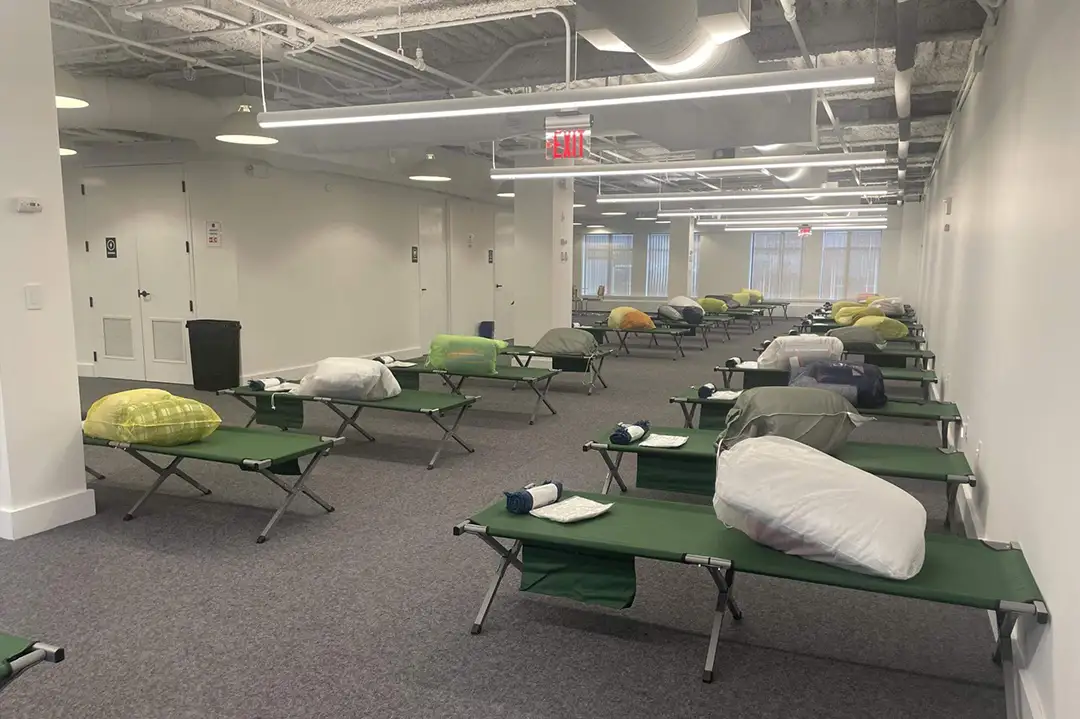 © 2024 Ted Resnikoff/UUA Rows of green cots in an open office space with white walls and gray carpet. The cots have bundles on them.